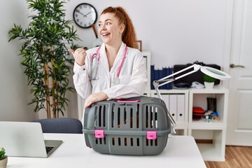 Canvas Print - Young redhead veterinarian woman working at pet clinic pointing thumb up to the side smiling happy with open mouth