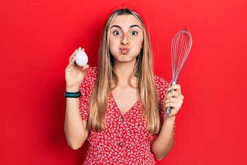 Poster - Beautiful hispanic woman holding egg and baker whisk puffing cheeks with funny face. mouth inflated with air, catching air.