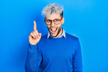 Wall Mural - Young hispanic man with modern dyed hair wearing sweater and glasses pointing finger up with successful idea. exited and happy. number one.