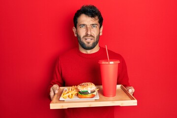 Poster - Handsome man with beard eating a tasty classic burger with fries and soda clueless and confused expression. doubt concept.