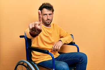 Canvas Print - Handsome man with beard sitting on wheelchair pointing with finger up and angry expression, showing no gesture