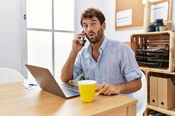 Young handsome man working at the office speaking on the phone scared and amazed with open mouth for surprise, disbelief face