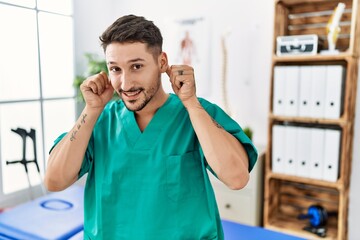 Canvas Print - Young physiotherapist man working at pain recovery clinic smiling pulling ears with fingers, funny gesture. audition problem