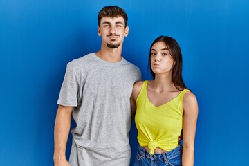 Poster - Young hispanic couple standing together over blue background puffing cheeks with funny face. mouth inflated with air, crazy expression.