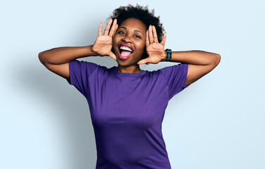 Wall Mural - African american woman with afro hair wearing casual purple t shirt smiling cheerful playing peek a boo with hands showing face. surprised and exited