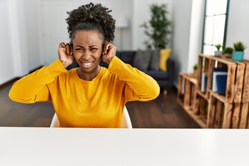 Sticker - Young african american woman wearing casual clothes sitting on the table at home covering ears with fingers with annoyed expression for the noise of loud music. deaf concept.