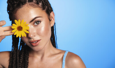 Beauty portrait of fashionable young woman with summer dreads hairstyle, tanned healthy facial skin, clear face, showing flower, standing over blue background