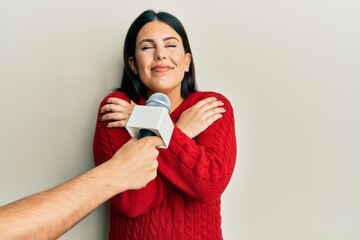 Poster - Beautiful brunette woman being interviewed by reporter holding microphone hugging oneself happy and positive, smiling confident. self love and self care