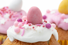 Close Up Of Easter Muffin With A White Glaze, Pink And Purple Decoration And Choloate Egg On Top, Another Two Similar Muffins Are On A Bokeh Background