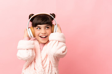 Wall Mural - Little caucasian girl isolated on pink background in pajamas and holding a pillow and listening music