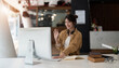 © Natee Meepian - Joyful young asian female in wireless headphones waving wand greeting to screen while sitting at table and having video chat with business partners using laptop against of comfortable office