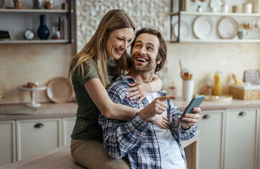 Cheerful millennial caucasian wife hugging husband with stubble and looking at smartphone in modern kitchen