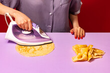 Creative Portrait Of Girl Ironing Pancakes On Lilac Color Tablecloth. Vintage, Retro Style Interior. Pop Art, Surrealism