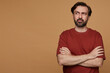 © timtimphoto - indoor portrait of bearded male posing over beige background wears red t-shirt, keeps his hands crossed on chest, looks aside thoughtfully, with confused facial expression