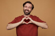 indoor portrait of bearded male posing over beige background shows heart sign with broad smile on his face