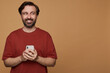 © timtimphoto - indoor portrait of bearded male posing over beige background smiles broadly, holding phone on his hand and looks aside