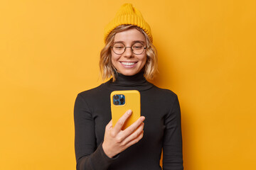 Wall Mural - Happy positive beautiful woman with fair hair holds smartphone broses social media content connected to internet downloads new application on new gadget wears yellow hat and black turtleneck