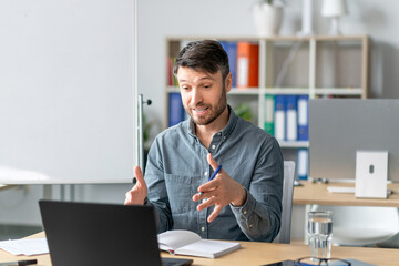 Mature male teacher looking at laptop and gesturing, sitting in office interior, explaining rules to student remotely