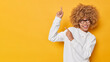 © WHstudio Leushin N - Cheerful curly haired adorable woman points above against blank space shows information banner demonstrates advertisement wears spectacles white casual jumper isolated over yellow background
