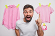 © WHstudio Leushin N - Amazed man gets excellent idea keeps index finger raised exclaims loudly realizes how to sell his old clothes poses against white background with plastered shirt t shirt and headphones behind