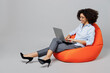 © ViDi Studio - Full body young employee business corporate lawyer woman of African American ethnicity in formal shirt work in office sit in bag chair hold use work on laptop pc computer isolated on grey background.