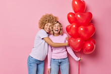 Two Positive Young Women Have Good Relationships Embrace Each Other With Love Hold Bunch Of Heart Helium Balloons Dressed In Casual Clothes Isolated Over Pink Background. Celebration Concept