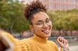 Portrait of attractive smiling curly haired young woman in optical round glasses poses for selfie outdoors walks in urban setting during leisure time photographs herself has free day or weekend