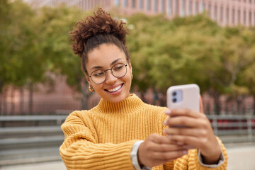 Wall Mural - Positive millennial content maker shoots influence video vlog enjoys networking lifestyle takes selfie via smartphone wears round eyeglasses and yellow jumper poses against blurred background