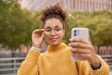 Wall Mural - Pleased confident woman with curly hair takes selfie pictures or shoots video for sharing to web blog keeps hand on rim of spectacles dressed in yellow knitted sweater poses at street outdoors