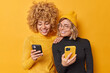 © wayhome.studio  - Beautiful two female friends use mobile phones browse internet type sms message dressed in casual turtlenecks smile gladfully isolated over vivid yellow background watch video via media app.