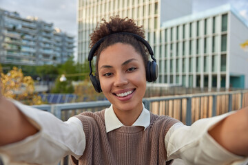 Wall Mural - Positive lovely teenage girl with combed curly hair takes selfie in urban setting smiles happily listens music from playlit wears wireless headphones on ears being in good mood during spring day