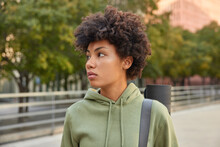 Active Healthy Woman With Curly Hair Dressed In Hoodie Carries Rolled Fitness Mat Concentrated Away Thoughtfully Strolls Outdoors During Sunny Day Returns Home After Training. Sport Concept.
