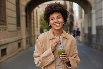 Wall Mural - Glad curly haired woman in casual jacket drinks drinks fresh detox vegetable smoothie keeps to healthy diet smiles happily strolls outside spends free time in city. People dieting lifestyle concept