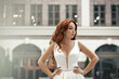 Refined feminine lady with red hair in a white dress. A happy and satisfied bride poses on a city street. Summer mood