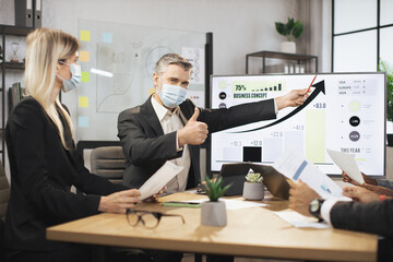 Wall Mural - Mature bearded chief in face mask, having a meeting together with his focused international colleagues, and showing business strategy on interactive digital screen with financial marketing charts.