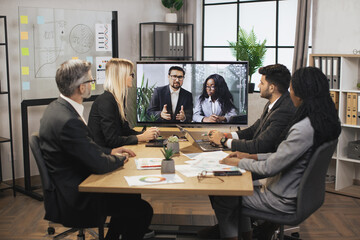 Wall Mural - Confident Caucasian business man and African woman tells business program concept to their multinational colleagues during a video conference in a modern office with a huge TV for conversations