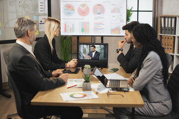 Wall Mural - Team of four focused multiethnical businesspeople looking at laptop, having video conference in modern office, with their male colleague explaining some financial charts. Telemeeting.