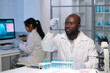 © pressmaster - African American male virologist in gloves and lab coat looking at blue liquid in flask against female colleague working in front of computer