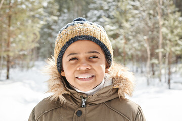 Cute contemporary multi-ethnic schoolboy in warm winterwear looking at camera against coniferous trees covered with snow