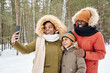 © pressmaster - Happy affectionate African American family in warm winterwear making selfie in park or forest while woman holding smartphone