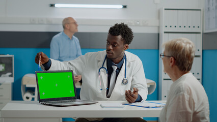 Wall Mural - Young medic having laptop with green screen technology on desk while talking to elder patient at healthcare clinic. Horizontal device with isolated mockup template and chroma key background