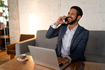 Wall Mural - Cheerful young businessman sitting on sofa and talking on cellphone in office.