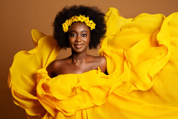 Wall Mural - Happy Dark Skinned Woman in Yellow Fashion Dress. Beauty Afro American Model dancing in Silk Gown waving Flying on Wind over Beige Background. Yellow Flower Wreath in Women Black Curly Hairstyle