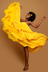 Wall Mural - Expressive Woman dancing in Yellow Flying Dress. Happy Dark Skinned Dancer in Waving Fabric Gown. Model with Black curly Afro Hair jumping over Beige Background