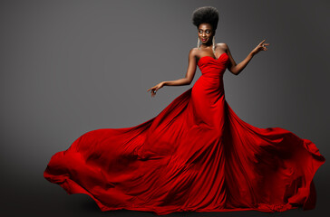 Wall Mural - Fashion Model in Red Long waving Luxury Dress. Dark Skinned Beauty Woman with Afro Black Hairstyle dancing over Gray Background. Happy African Sexy Girl in Silk Evening Gown with flying Fabric