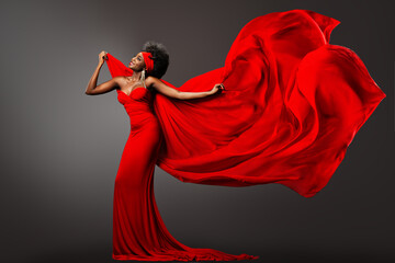 Fashion African Woman in Red Dress with Silk Scarf flying on Wind. Happy Dark Skinned Model Dancing with Fluttering Chiffon Fabric over Gray Background. Afro Female in Long Gown with waving Cloth