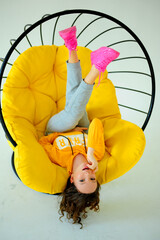 Wall Mural - a girl in a bright yellow sweatshirt sits on a juicy yellow chair and writhes faces different emotions on the white background of the studio teenager is having fun and she is smiling.