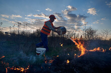 Firefighter Ecologist Fighting Wildfire In Field With Evening Sky On Background. Male Environmentalist Holding Bucket And Pouring Water On Burning Dry Grass. Natural Disaster Concept.