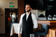 © Andrii - A young waiter with a beard puts on an apron and prepares for a working day in a fine restaurant.