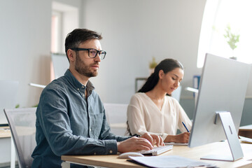 Focused Caucasian man using computer, working in modern office with his colleague, copy space
